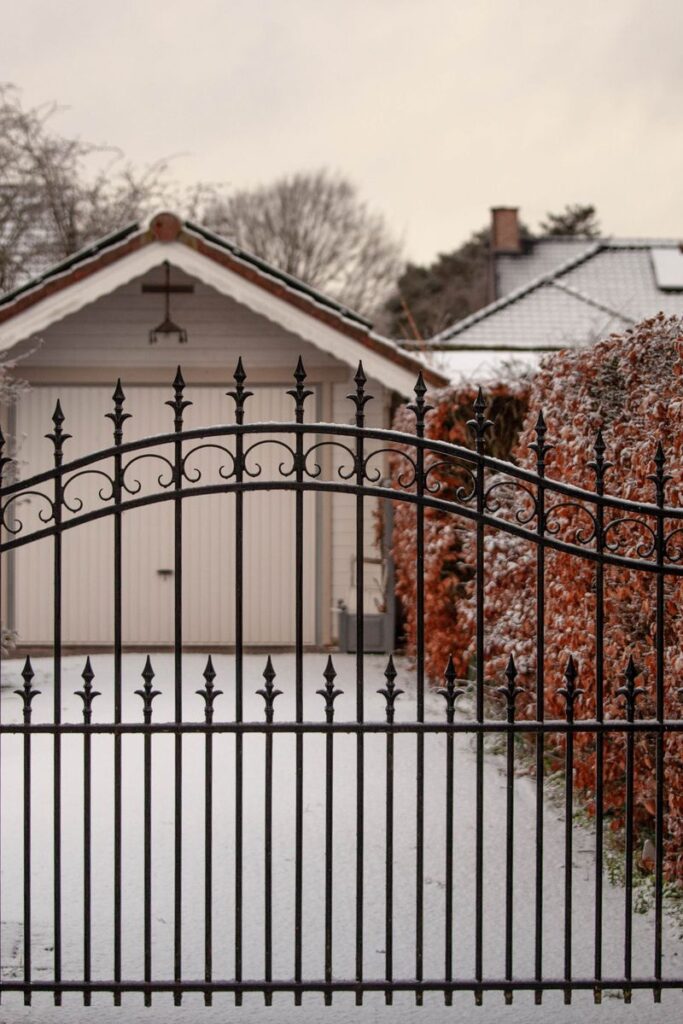 A serene snowy courtyard featuring a decorative iron gate and surrounding hedges in winter.